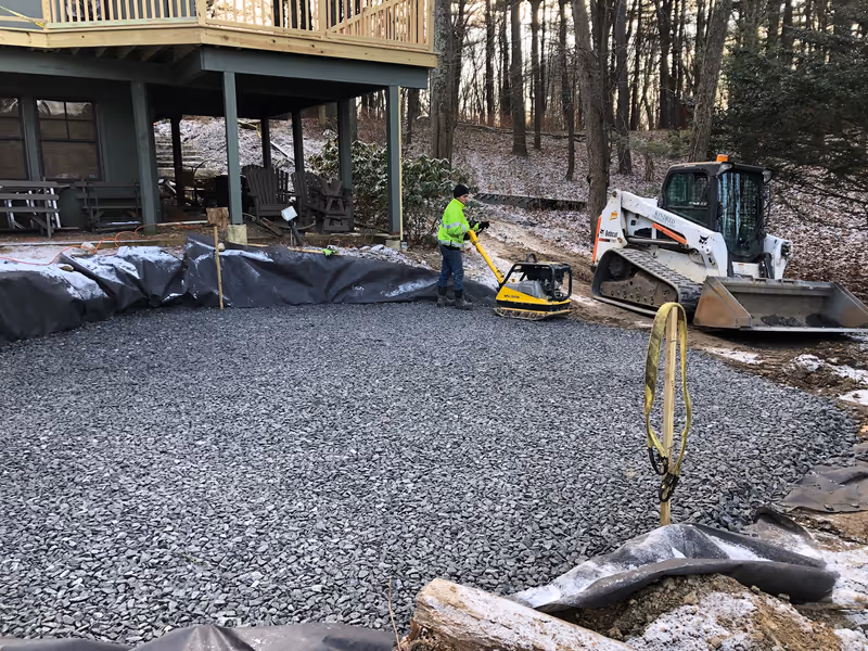 Crew member compacting aggregate beside a house with a tracked loader nearby.