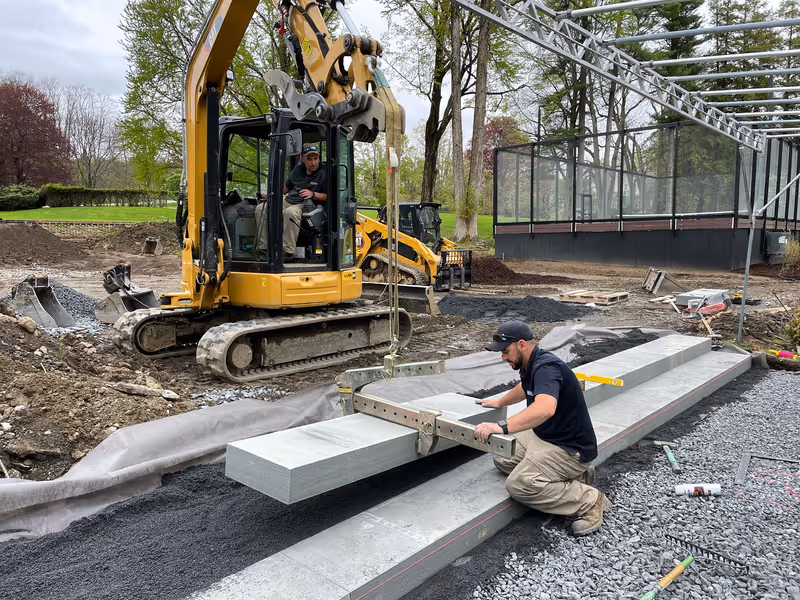 Crew positioning a large stone step with an excavator and lifting clamp beside a pool terrace.