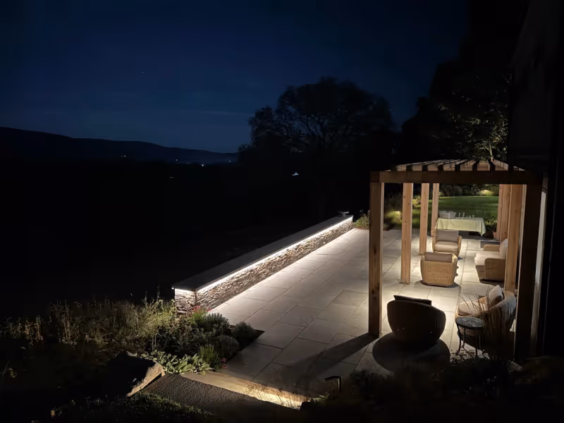 Illuminated stone wall along a terrace and covered seating area at night.