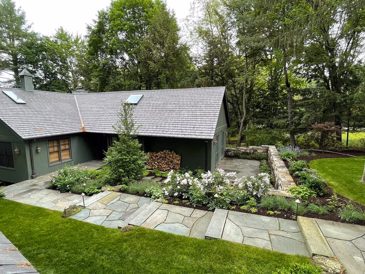 Finished stone paths, steps, planted beds and retaining wall around a wooded residence.